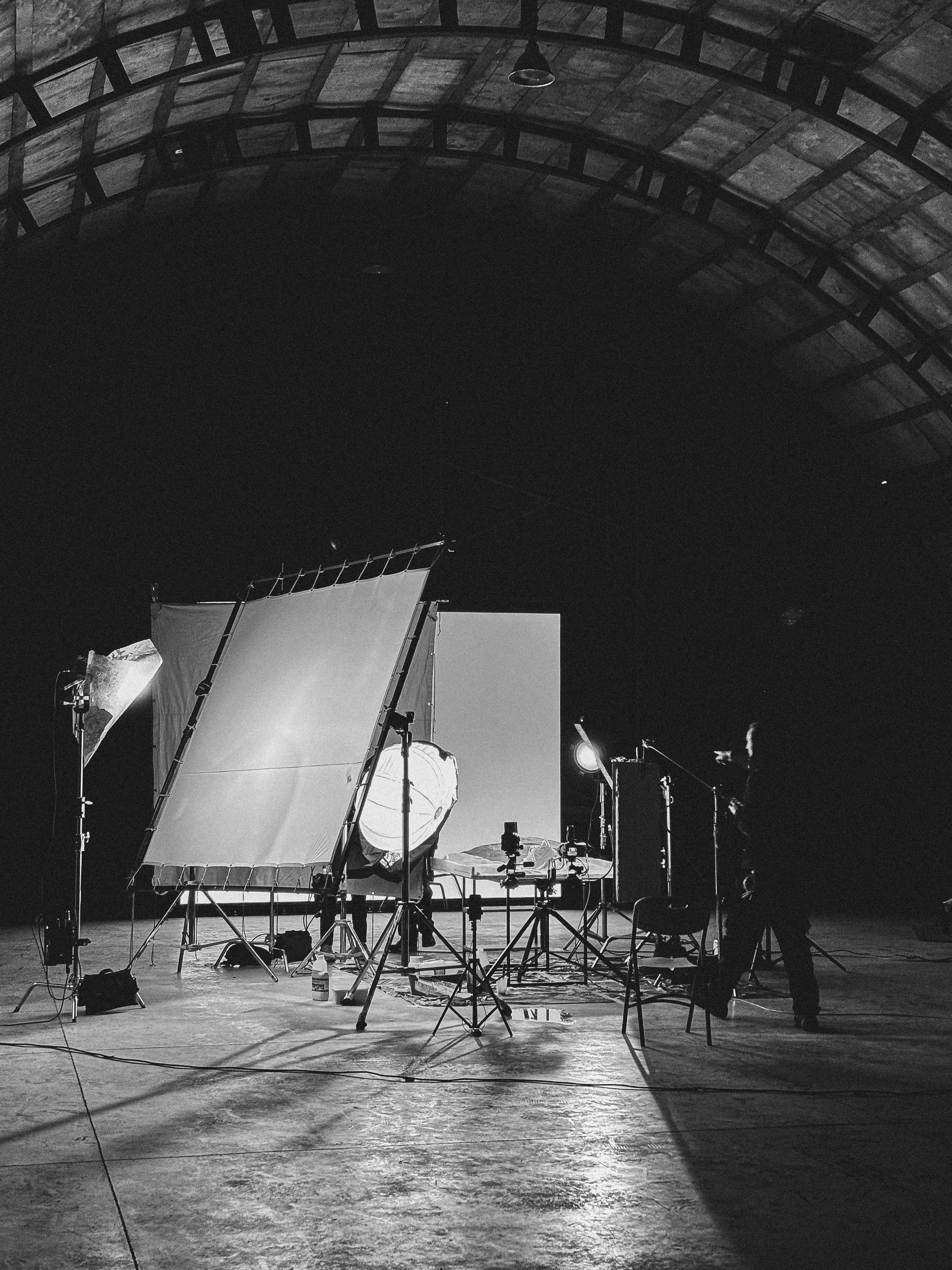 Black and white photo of a dimly lit film studio with lighting equipment under a large arched ceiling.