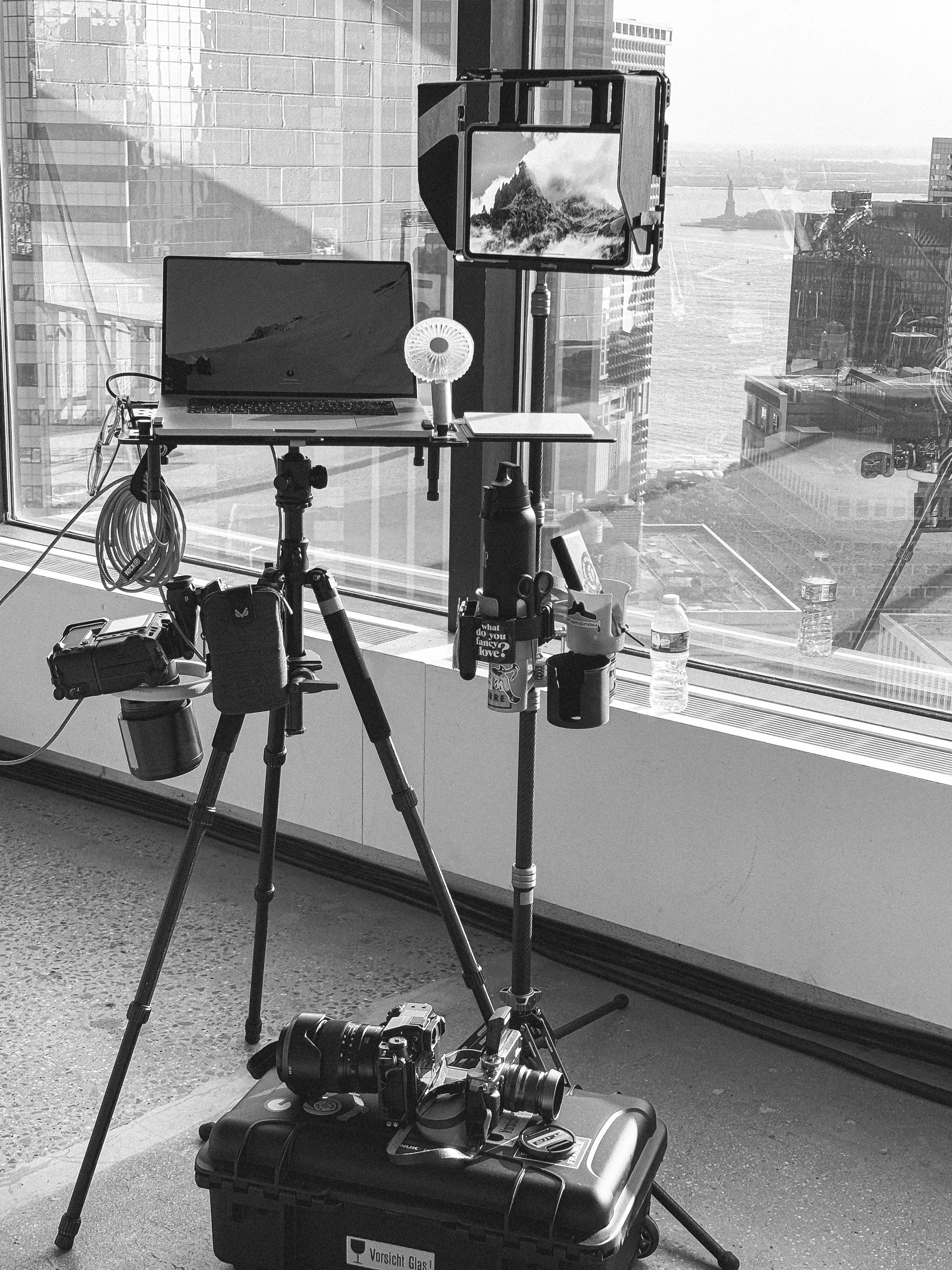 Black and white photo of a photography setup with laptop and cameras, Statue of Liberty visible through windows.